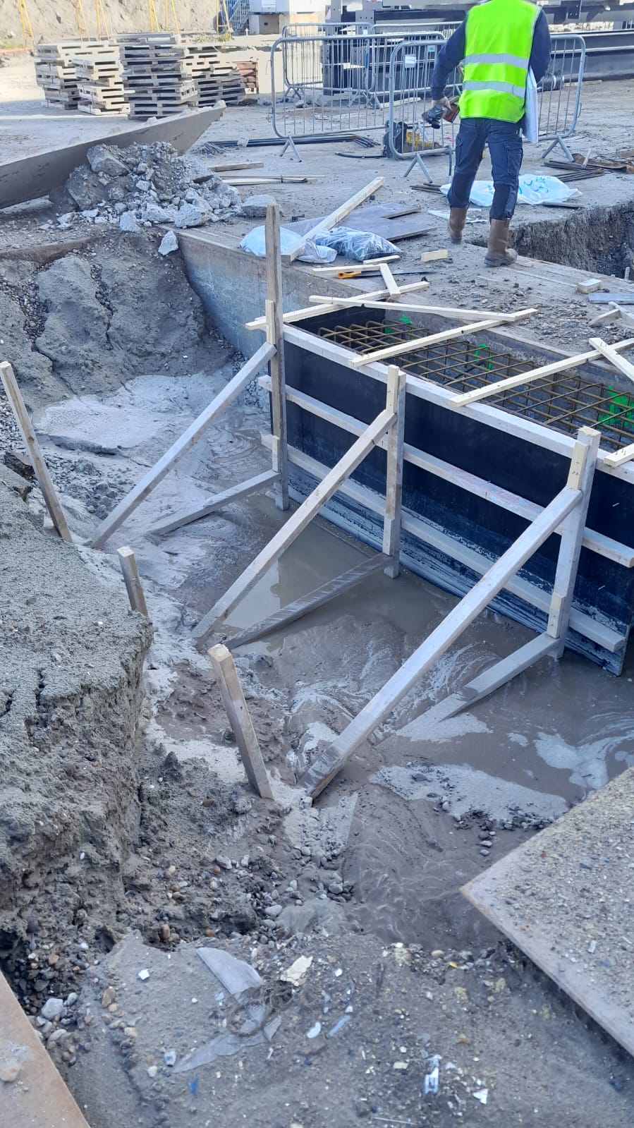 Bouwplaats met betonnen bekisting in de grond en een werker in veiligheidshesje.